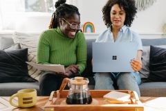 Two colleagues sit on a couch with a laptop and papers. A tray with coffee is on the table in front of them. Opens media file when clicked. 